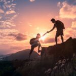 Couple hiking together and helping each other climb a mountain at sunset, representing progress toward financial goals.