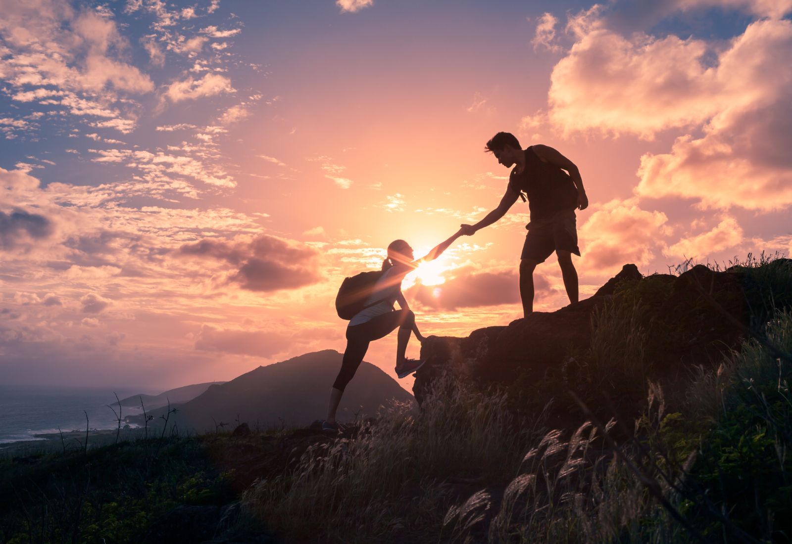 Couple hiking together and helping each other climb a mountain at sunset, representing progress toward financial goals.